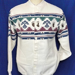 Könnte beinhalten: Ein weißer, geknöpfter Pullover mit einem indianisch inspirierten Muster in Blautönen, Grün, Rot und Braun. Das Muster zeigt stilisierte Vögel und Bärenpfotenabdrücke.