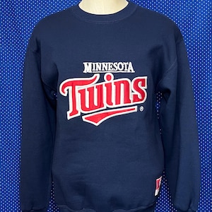 Può includere: Una felpa blu navy con il logo dei Minnesota Twins in lettere rosse e bianche.