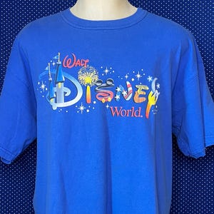 May include: A blue t-shirt featuring the "Walt Disney World" logo in a colorful design. The graphic includes a castle, fireworks, and the word "Disney" in red, white, and blue. The shirt is short-sleeved and made of a soft material.
