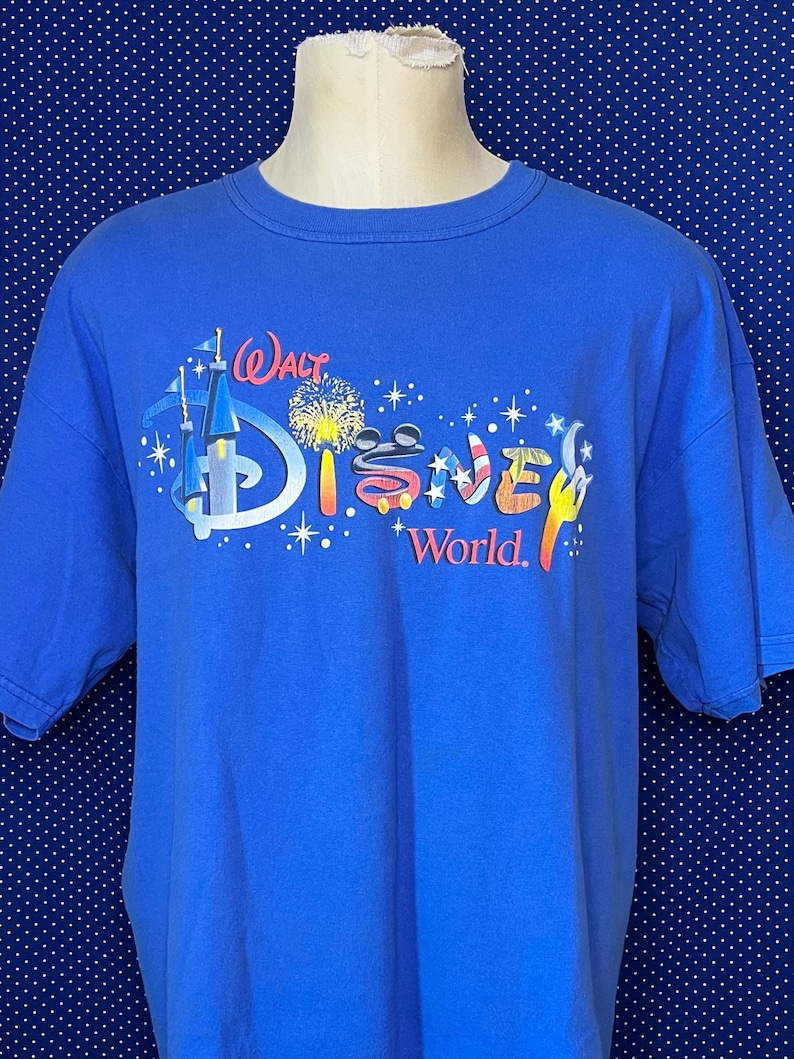 May include: A blue t-shirt featuring the "Walt Disney World" logo in a colorful design. The graphic includes a castle, fireworks, and the word "Disney" in red, white, and blue. The shirt is short-sleeved and made of a soft material.