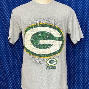 Puede incluir: Camiseta gris con un logotipo verde y blanco de los Green Bay Packers. El logotipo es un collage de imágenes relacionadas con el equipo. El texto "Packers" está debajo del logotipo.