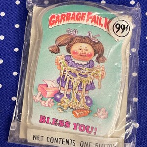 May include: A plastic-wrapped button featuring a cartoon image of a Cabbage Patch Kid doll covered in green slime. The text "Garbage Fail Kid" and "Bless You!" are printed on the button. The button is priced at 99 cents.