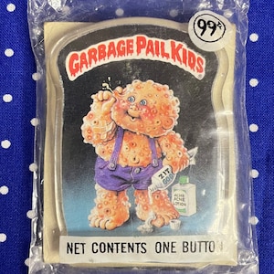 May include: A Garbage Pail Kids button featuring a green-skinned character with a large zit on their nose. The character is wearing blue overalls and holding a tube of acne lotion. The button is packaged in a plastic bag with a price tag of 99 cents.