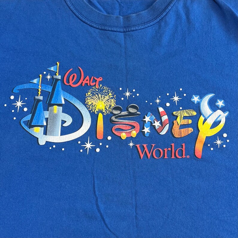 May include: Blue t-shirt featuring the Walt Disney World logo. The logo includes the word "Walt" above a stylized "Disney" with fireworks and a castle graphic. The words "World" are below the Disney logo.