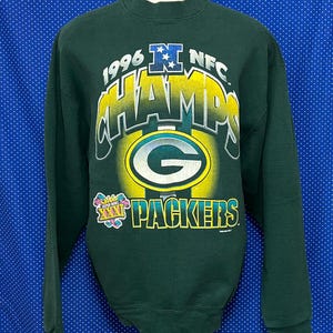 Puede incluir: Sudadera verde con el texto "1996 NFC Champs" y una "G" verde con contorno amarillo. El texto "Packers" está debajo de la "G" y un logotipo de Super Bowl XXXI está a la izquierda del texto.