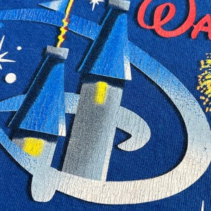 May include: Close-up of a blue t-shirt featuring a graphic of a castle with yellow and blue accents, and the word "Walt" in red script. The design includes a stylized "D" logo and starbursts.