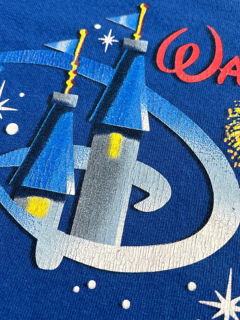 May include: Close-up of a blue t-shirt featuring a graphic of a castle with yellow and blue accents, and the word "Walt" in red script. The design includes a stylized "D" logo and starbursts.