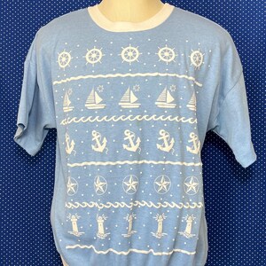 May include: A light blue short-sleeved t-shirt with a white nautical design. The design features anchors, sailboats, stars, lighthouses, and ship's wheels.