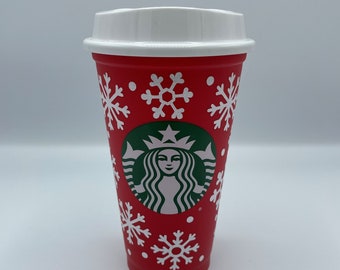 May include: A red Starbucks holiday cup with a white lid. The cup features white snowflake designs and the Starbucks logo in green. The cup is designed for hot beverages.