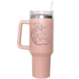 May include: A pink stainless steel travel mug with a handle and a straw. The mug features a design of an apple with a stem and leaf, and the text "Mrs. Ramsey" inside the apple.