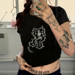 May include: Black crop top featuring a white graphic of a skeleton skateboarding, surrounded by a lightning bolt design. The image also shows a tattoo that says "END OF TIME" in gothic lettering.