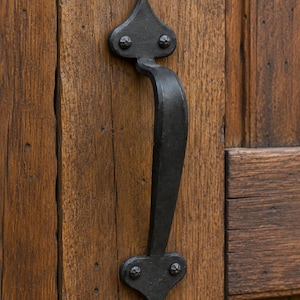 May include: A black, wrought iron door handle with a curved design, mounted on a weathered, wooden door. The handle has decorative, heart-shaped ends and is secured with rivets. The wood grain is visible.