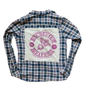 May include: A blue and white plaid flannel shirt with a gray patch on the back. The patch features a purple logo that says "Visit Historic Schrute Farms, Beet Winery, Pa."
