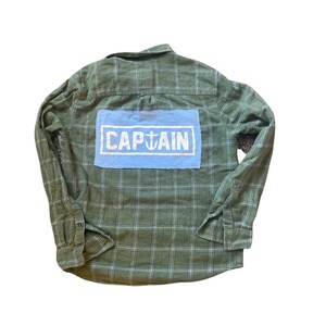 May include: A green and white plaid flannel shirt with a blue patch on the back that says "CAPTAIN" in white letters with an anchor symbol.