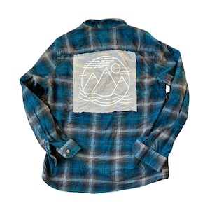 May include: A blue and black plaid flannel shirt with a white embroidered patch on the back depicting mountains, a sun, and waves.