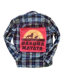 Może przedstawiać: Koszula flanelowa w niebiesko-czerwoną kratę z czerwoną, żółtą i pomarańczową łatą z tyłu. Łata przedstawia zachód słońca z lwem i ptakiem oraz tekstem "Hakuna Matata".