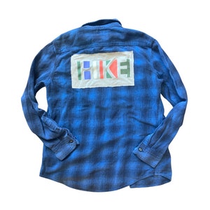 Könnte beinhalten: Ein blaues Flanellhemd mit Karomuster und einem weißen Patch auf der Rückseite, auf dem "HIKE" in bunten Buchstaben steht.