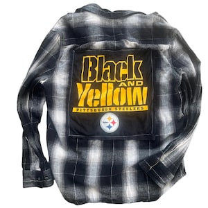 May include: A black and white plaid long-sleeve button-down shirt featuring a large black rectangular patch on the back. The patch displays the yellow text Black and Yellow, Pittsburgh Steelers, and a circular Pittsburgh Steelers team logo.
