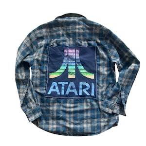 May include: Blue and gray plaid flannel shirt with a dark blue patch featuring the Atari logo in a pixelated rainbow design. The word "ATARI" is printed below the logo in blue letters.