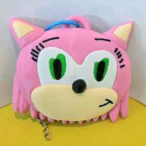May include: A pink, 3D-printed keychain featuring a character with green eyes, a black nose, and a light yellow muzzle. The character has a blue loop at the top and a silver chain at the bottom. The character's face has a smiling expression.