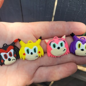 May include: Four colorful 3D printed character heads. The characters are yellow with green eyes, pink with green eyes, purple with yellow eyes, and black with red eyes.
