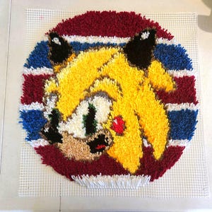 Peut inclure: Un tapis rond fait à la main représentant un personnage de dessin animé avec une fourrure jaune, des oreilles noires et un cœur rouge. Le tapis a une bordure rayée rouge, blanche et bleue, et est fait de fil sur un support en maille blanche.