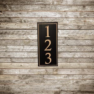 May include: A rectangular black address plaque with the numbers 1, 2, and 3 in a light brown color. The numbers are carved into the plaque, which has a light brown border. The background is a wooden wall.
