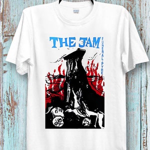 Puede incluir: Camiseta blanca con un estampado gráfico en blanco y negro de una figura esquelética con llamas y calaveras. El texto "THE JAM FUNERAL PYRE" está impreso en azul en la camiseta.