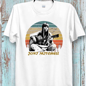 Puede incluir: Camiseta blanca con un gráfico de estilo vintage de Joni Mitchell tocando la guitarra frente a una puesta de sol. El texto "Joni Mitchell" está impreso debajo de la imagen.