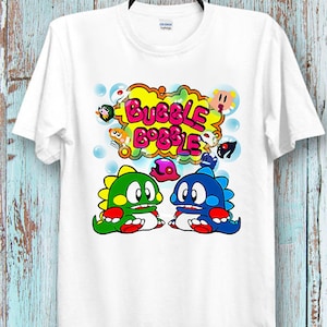 Op de afbeelding: Wit T-shirt met een kleurrijke afbeelding van het videospel Bubble Bobble. De afbeelding toont twee bellenblazende dinosaurussen, een spreekballon met de titel van het spel en andere kleurrijke personages.