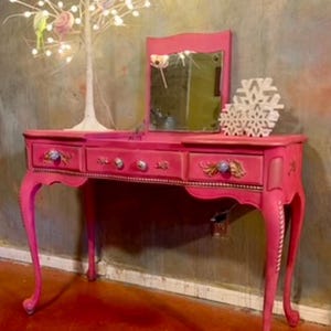 May include: A pink vanity table with three drawers and a mirror. The table has ornate legs and decorative details. A white twig tree with lights is in the background.