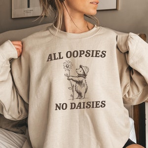 May include: A beige sweatshirt with a graphic of a bear holding a daisy. The text "All oopsies no daisies" is printed on the sweatshirt.