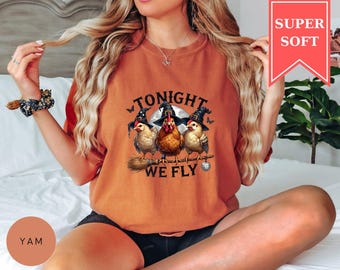 May include: A burnt orange crewneck sweatshirt featuring a graphic of three chickens wearing black witch hats, perched on a broomstick. The text reads TONIGHT WE FLY. The design includes a full moon, small butterflies, and a disco ball hanging from the broom.