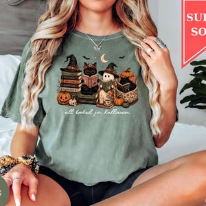 A woman wearing a green t-shirt with a Halloween-themed graphic of cats and books, sitting on a bed.