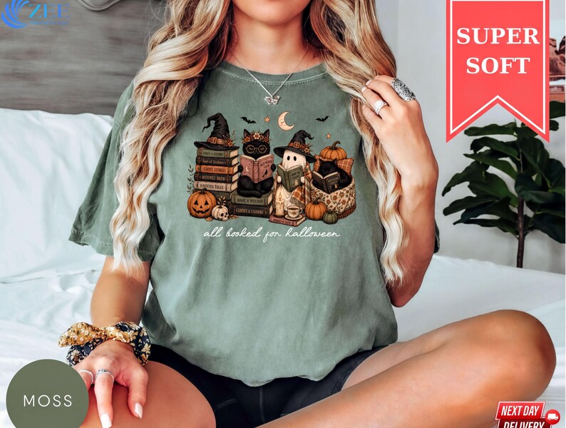 A woman wearing a green t-shirt with a Halloween-themed graphic of cats and books, sitting on a bed.