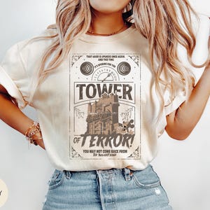 May include: Ivory-colored t-shirt featuring a vintage-style graphic for the Tower of Terror ride. The design includes the ride's name, a spooky building illustration, and the text "You may not come back from The Twilight Zone."