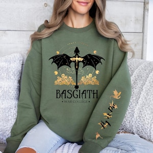 May include: An olive green crewneck sweatshirt featuring a black and gold dragon graphic on the chest with the text BASGIATH WAR COLLEGE. The left sleeve displays small gold dragon silhouettes and the text Fly Or Die in a vertical script font.