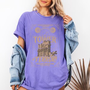 May include: A purple t-shirt featuring a graphic of the Tower of Terror ride. The design includes the words "Tower of Terror" in a stylized font, with additional text and decorative elements. The shirt is a comfortable, casual style.