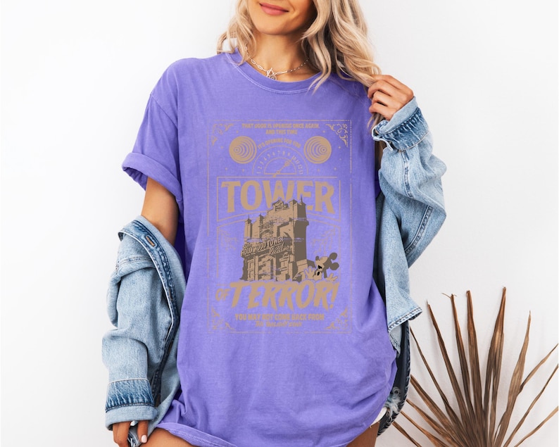 May include: A purple t-shirt featuring a graphic of the Tower of Terror ride. The design includes the words "Tower of Terror" in a stylized font, with additional text and decorative elements. The shirt is a comfortable, casual style.