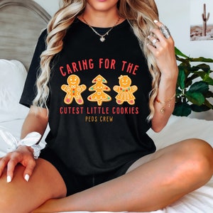 以下が含まれることがあります： 赤と白の文字「Caring for the Cutest Little Cookies Peds Crew」が書かれた黒いTシャツ。文字の周りを、ジンジャーブレッドマン、ジンジャーブレッドウーマン、ジンジャーブレッドツリーの3つのジンジャーブレッドクッキーが囲んでいます。