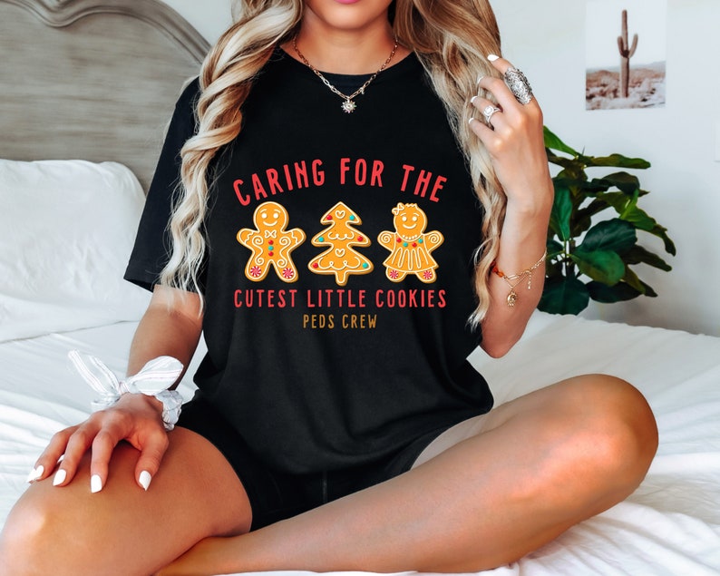 以下が含まれることがあります： 赤と白の文字「Caring for the Cutest Little Cookies Peds Crew」が書かれた黒いTシャツ。文字の周りを、ジンジャーブレッドマン、ジンジャーブレッドウーマン、ジンジャーブレッドツリーの3つのジンジャーブレッドクッキーが囲んでいます。