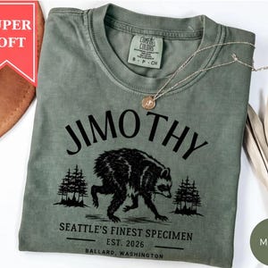 A green t-shirt with a graphic design of a bear and trees, along with text that says "Jimothy" and "SEATTLE'S FINEST SPECIMEN EST. 2026 BALLARD, WASHINGTON".