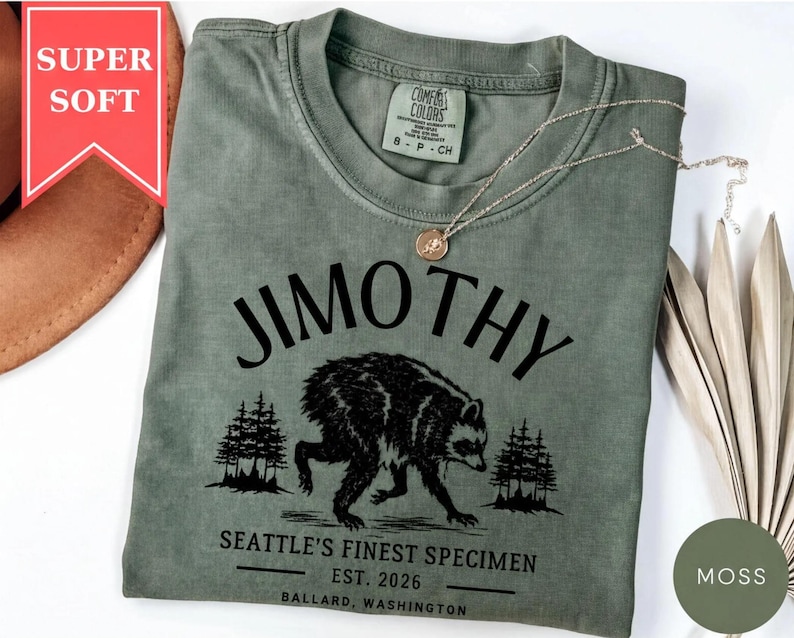A green t-shirt with a graphic design of a bear and trees, along with text that says "Jimothy" and "SEATTLE'S FINEST SPECIMEN EST. 2026 BALLARD, WASHINGTON".