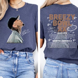 May include: Navy blue t-shirt featuring a portrait of a man wearing a black cap and denim jacket. The back of the shirt displays the text "Breezy Bowl XX Stadium World Tour" with a list of European and North American tour dates.