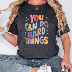 May include: A dark gray t-shirt with the text "YOU CAN DO HARD THINGS" in colorful letters and small hearts.