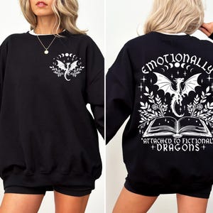 May include: Black sweatshirt with a white graphic print of a dragon, moon phases, and stars. The text on the back reads "Emotionally attached to fictional dragons."