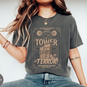 May include: Dark gray t-shirt with a distressed graphic featuring the text "TOWER OF TERROR!" and a vintage-style illustration. The shirt has a classic crew neck and short sleeves. The text "YOU MAY NOT COME BACK FROM THE TWILIGHT ZONE" is also visible.