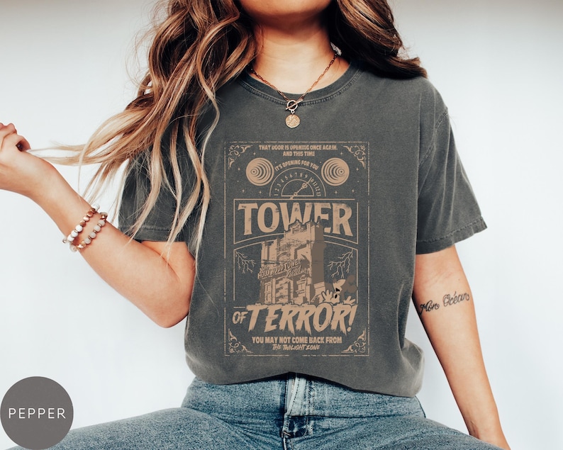 May include: Dark gray t-shirt with a distressed graphic featuring the text "TOWER OF TERROR!" and a vintage-style illustration. The shirt has a classic crew neck and short sleeves. The text "YOU MAY NOT COME BACK FROM THE TWILIGHT ZONE" is also visible.