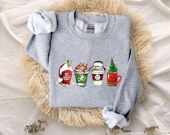 Puede incluir: Una sudadera gris claro de cuello redondo con un estampado gráfico de cuatro tazas con temática navideña. Las tazas muestran diseños festivos, incluyendo textos que dicen Are You Serious Clark, Merry Christmas y Full Lot Of Sap.