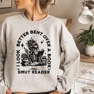 May include: A light gray crewneck sweatshirt featuring a black graphic print of a skeleton reading a book. The text reads I LOOK BETTER BENT OVER A BOOK and PROUD SMUT READER, surrounded by illustrations of stacked books, flowers, and stars.